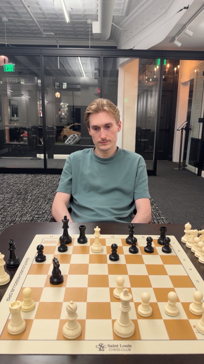 Cam in a teal t-shirt sitting at a chess board at the Saint Louis Chess Club, staring at the pieces with great concern