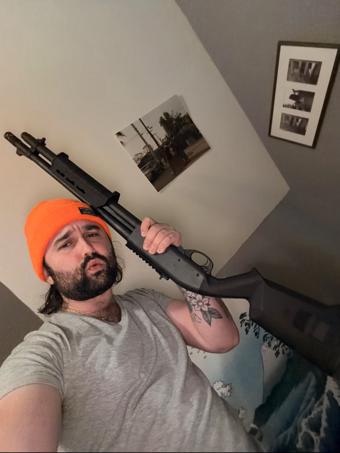 Toby in a bright orange beanie and grey t-shirt, posing with a pump-action shotgun over his shoulder in a bedroom with a wave tapestry